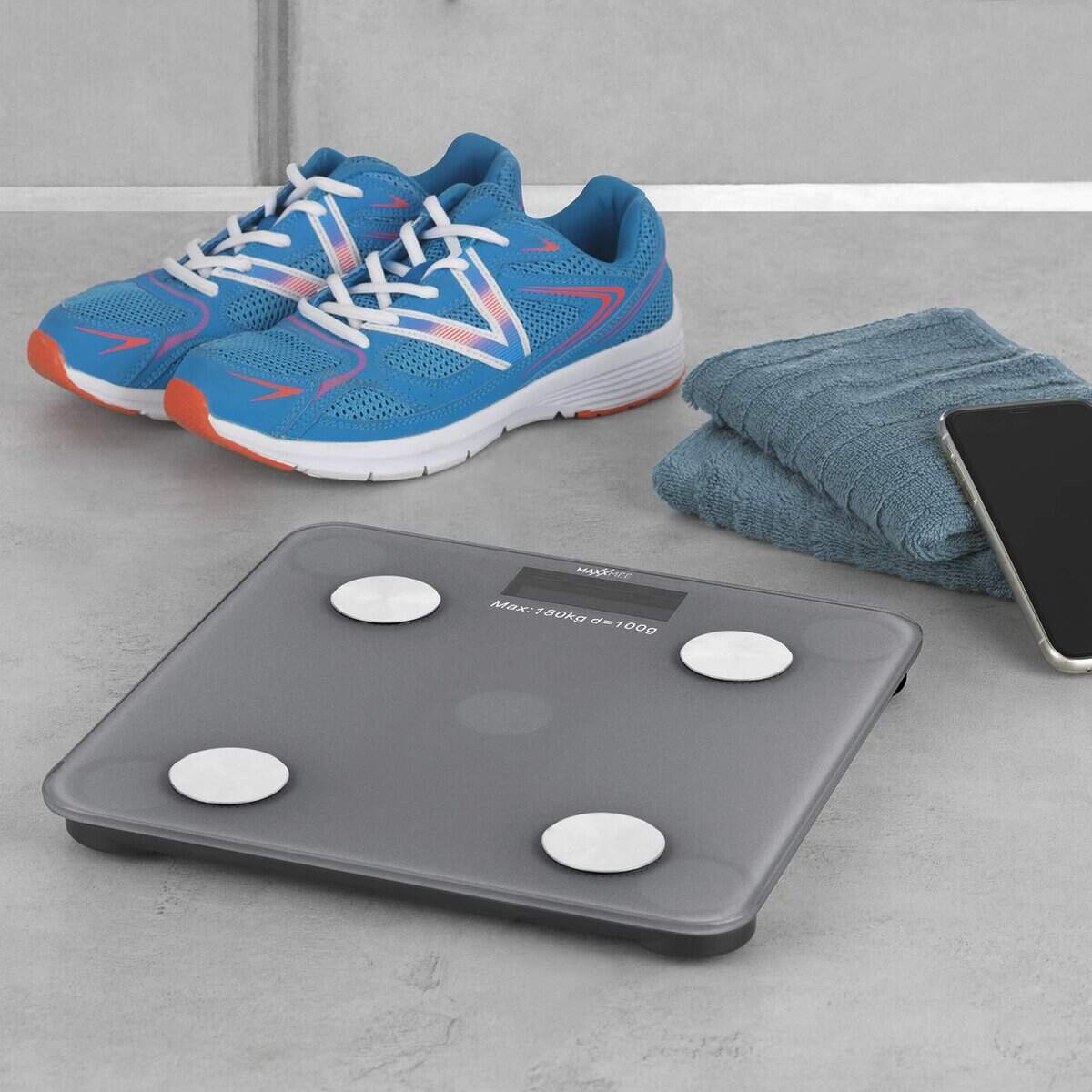 Ein Paar blaue Sportschuhe, ein gefaltetes Handtuch, ein Smartphone und eine digitale Personenwaage liegen auf dem grauen Boden.
